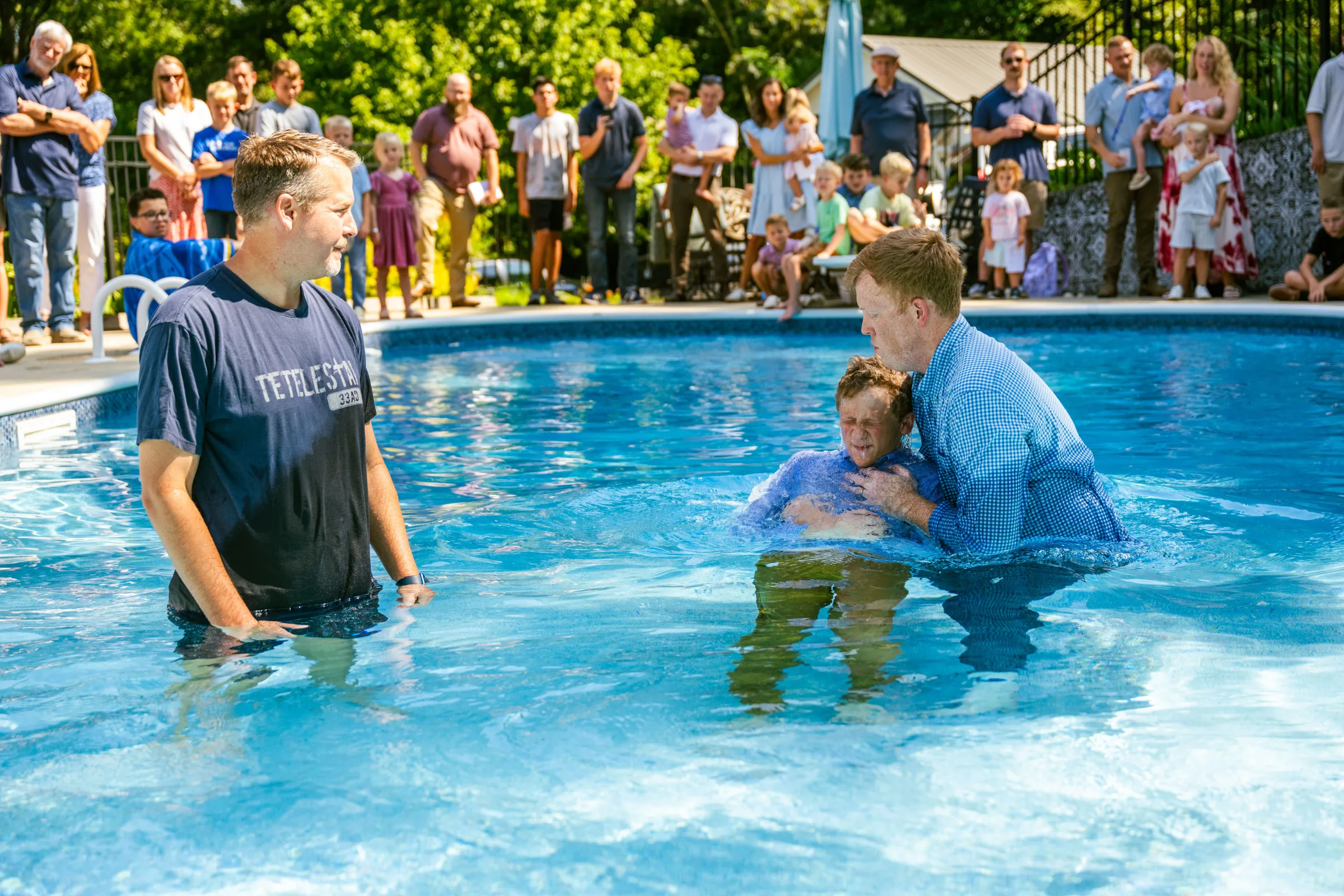 A child is baptized in a pool while Trinity Fellowship members gather around to watch.